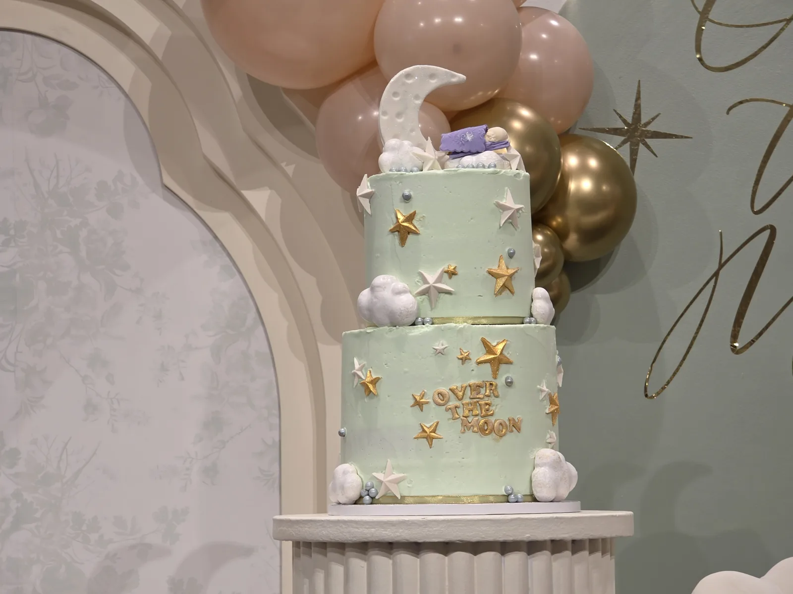 A two tier pale mint green cake decorated with white and gold stars, fluffy clouds, silver pearls, a crescent moon topper, and gold Over The Moon lettering.