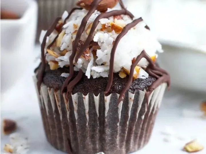Chocolate cupcake in a dark liner topped with white frosting, chopped nuts, and a chocolate drizzle.