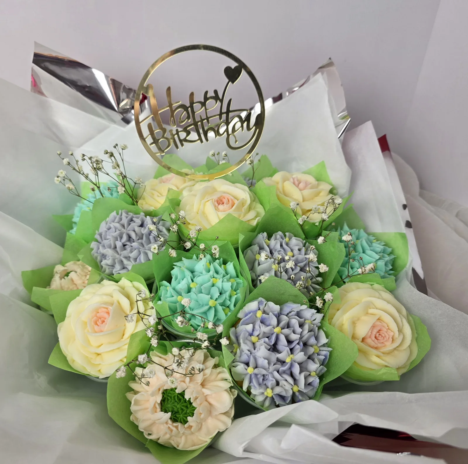 A bouquet of cupcakes with buttercream roses, hydrangeas and blooms in cream, lavender, mint and peach, nestled in green tissue with a gold Happy Birthday topper.