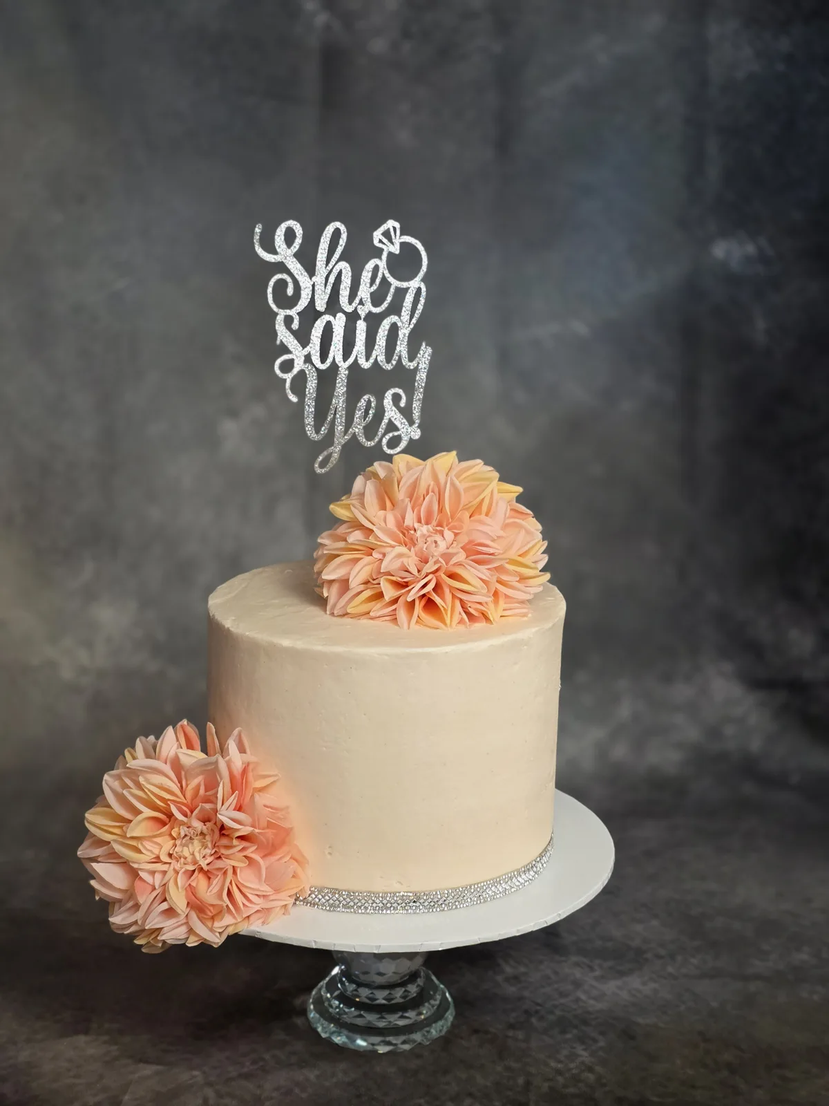 A single tier buttercream wedding cake in pale peach, adorned with two large coral and peach dahlia sugar flowers and a silver rhinestone base trim.