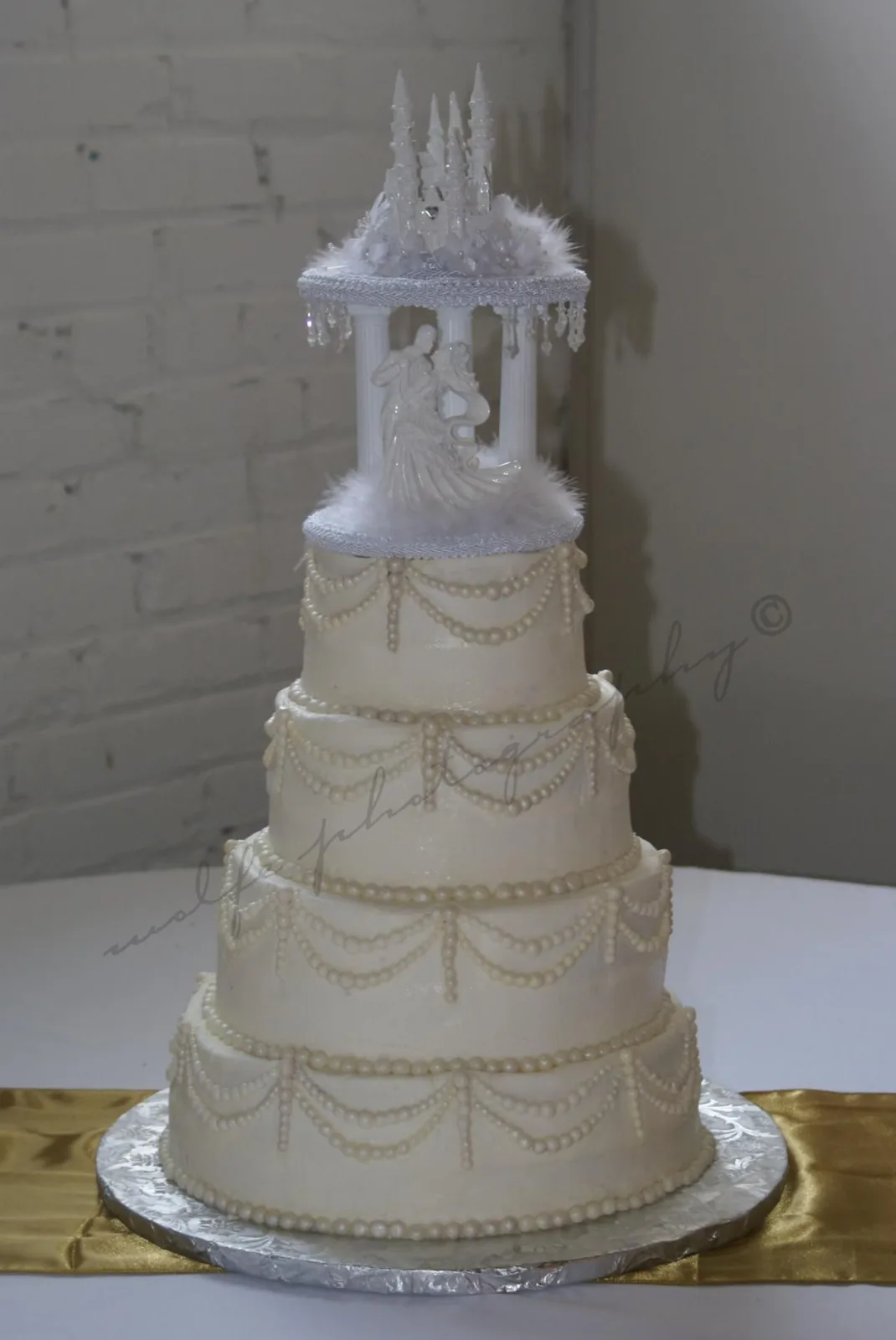 Tall four tier white wedding cake with draped pearl swags on each tier, topped with a white gazebo featuring a bride and groom figurine inside.