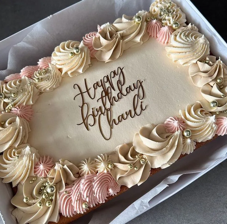 Rectangular sheet cake with cream frosting, piped pink and ivory rosettes around the edges, gold and silver sugar pearls, and a dark chocolate Happy Birthday message.