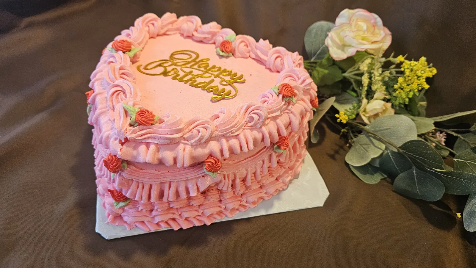 Heart shaped pink frosted cake with ruffled side layers, piped rose rosettes, small orange rosebuds with green leaves, and gold Happy Birthday script on top.