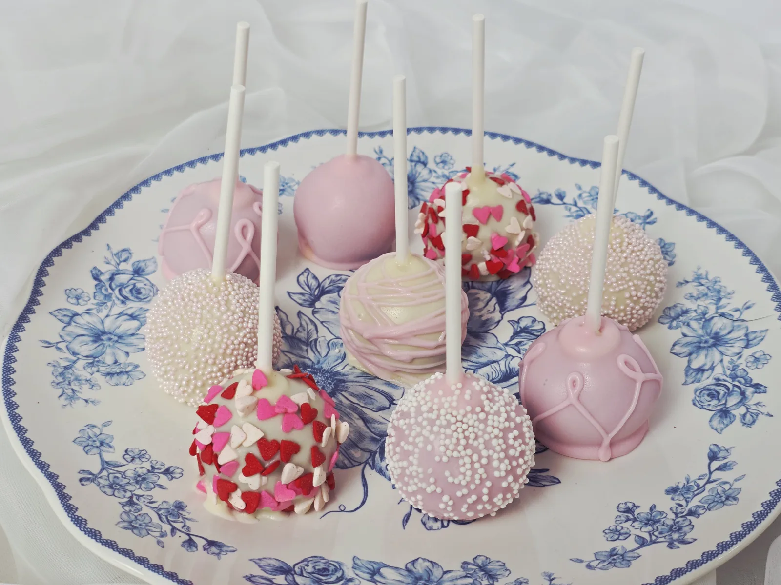 Round cake pops in pink and white chocolate decorated with heart sprinkles, nonpareils, and drizzle patterns arranged on a blue floral plate.