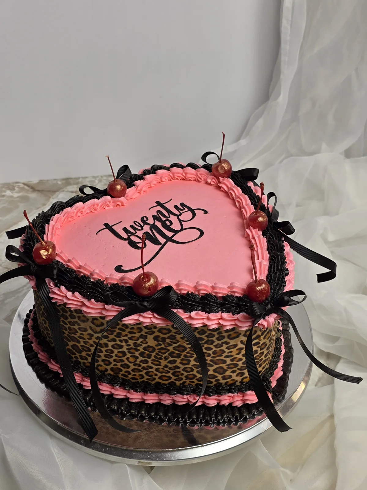 Round cake featuring a pink frosted top with black script writing, surrounded by red cherries and black ribbon bows over pink and black ruffled borders with leopard print sides.