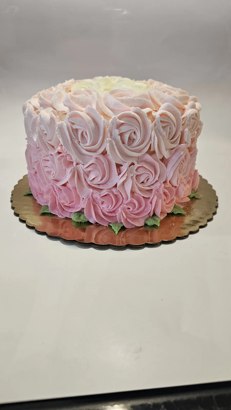 Round cake entirely covered in buttercream rosettes that ombre from cream at the top to soft pink at the base, accented with green leaves on a scalloped rose gold board.