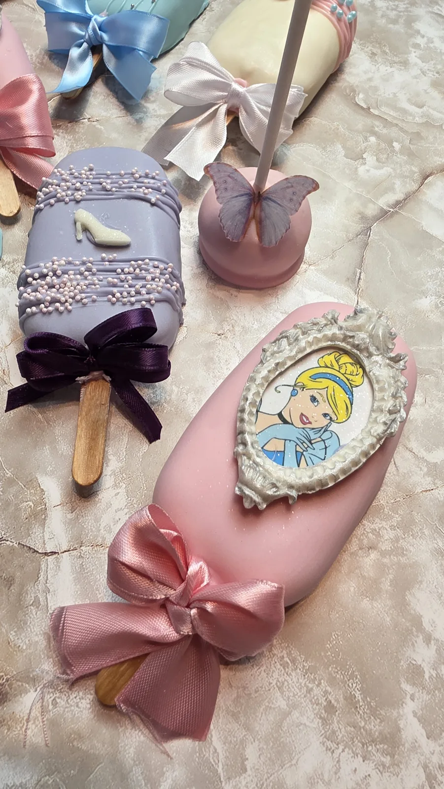 Pink chocolate coated cakesicle featuring a blonde princess portrait in an ornate silver frame, finished with a large pink satin bow on a wooden stick.