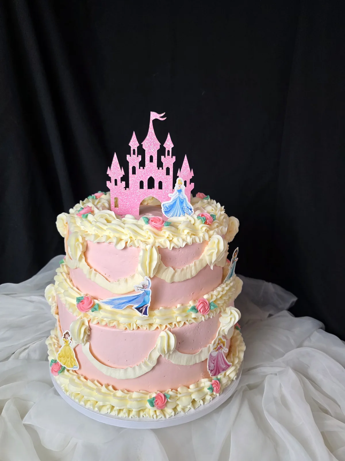 Three tiered pink frosted cake decorated with cream piped swags, pink buttercream roses, princess figurine cutouts, and topped with a pink glitter castle.