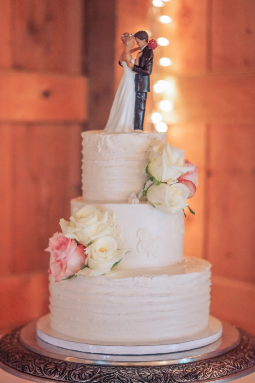 Three tier white buttercream wedding cake with rustic textured frosting topped with a bride and groom figurine and decorated with white and blush pink roses.