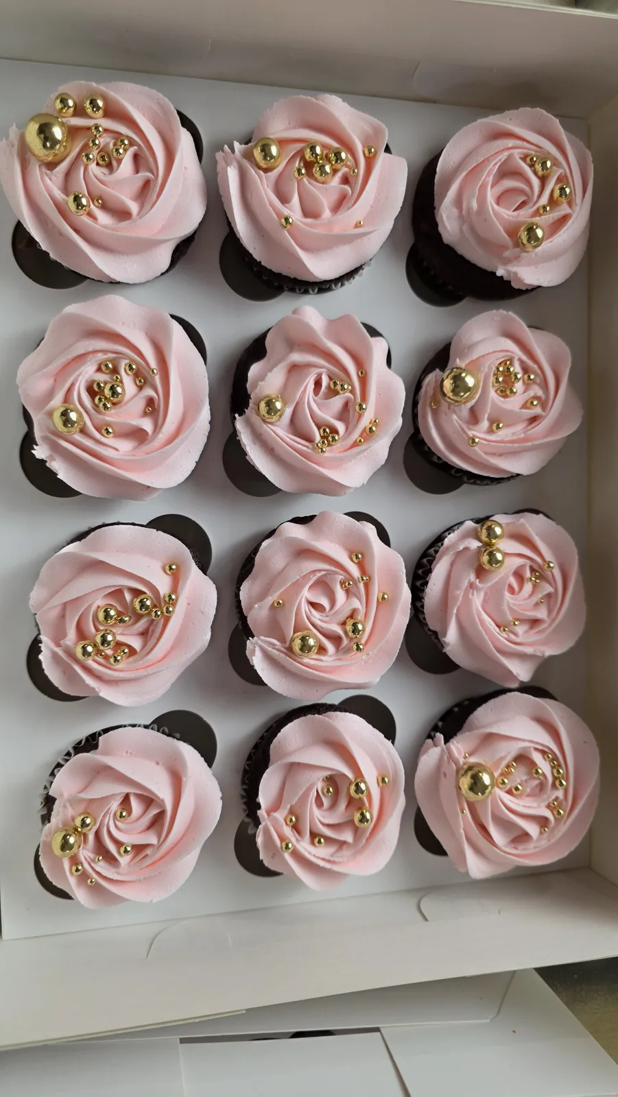 Twelve cupcakes arranged in a white box, each topped with pale pink buttercream piped into rose shapes and decorated with metallic gold pearl accents.