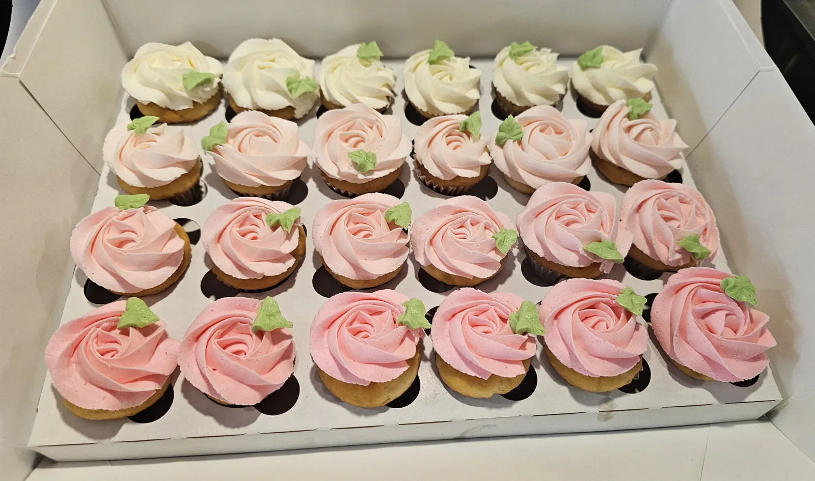 Box of vanilla cupcakes topped with rose shaped buttercream swirls in white, pale pink, and deeper pink shades with small green fondant leaves.