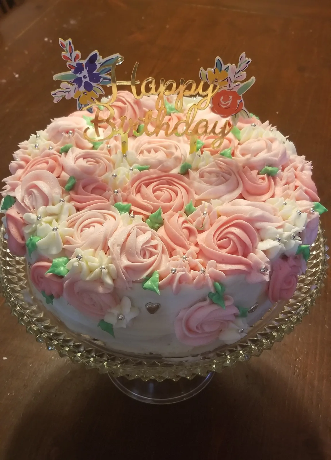 A round frosted cake covered in piped pink and white buttercream roses with green leaves, silver dragees, and a gold Happy Birthday floral topper on a crystal glass stand.