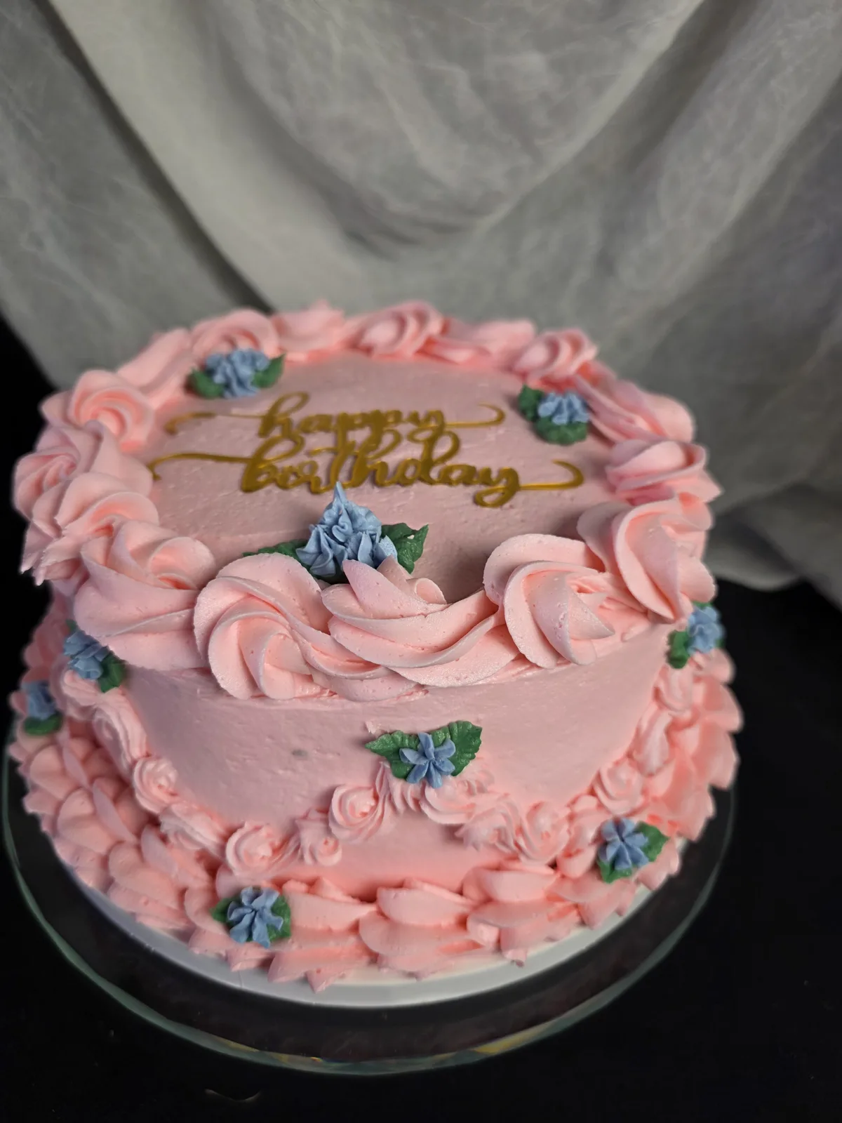 A pink frosted birthday cake decorated with piped buttercream rosettes, small blue floral accents with green leaves, and a gold happy birthday script on top.