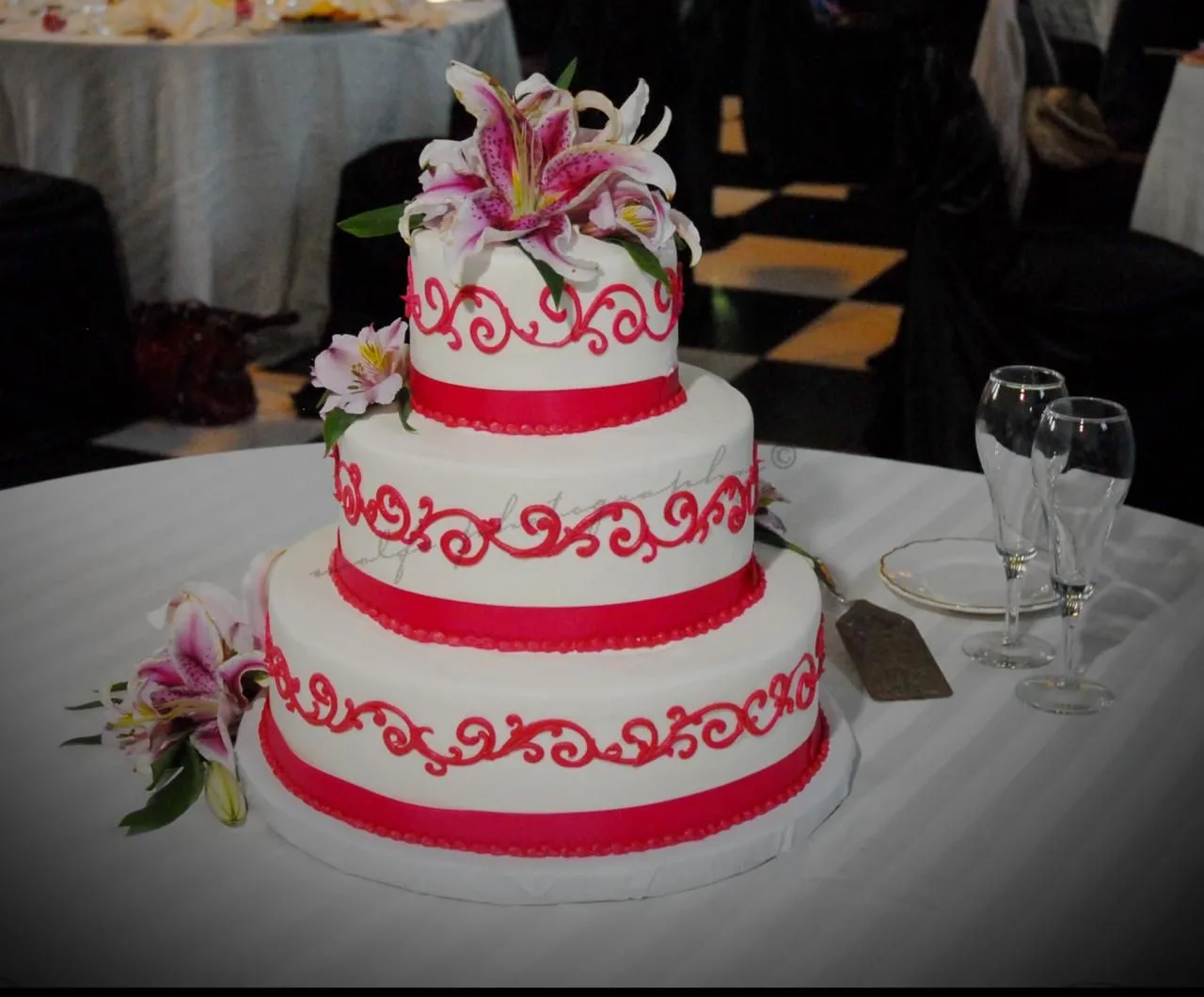 Three tier white cake with hot pink filigree scrollwork and ribbon trim, topped with fresh pink and white stargazer lilies cascading down the sides.