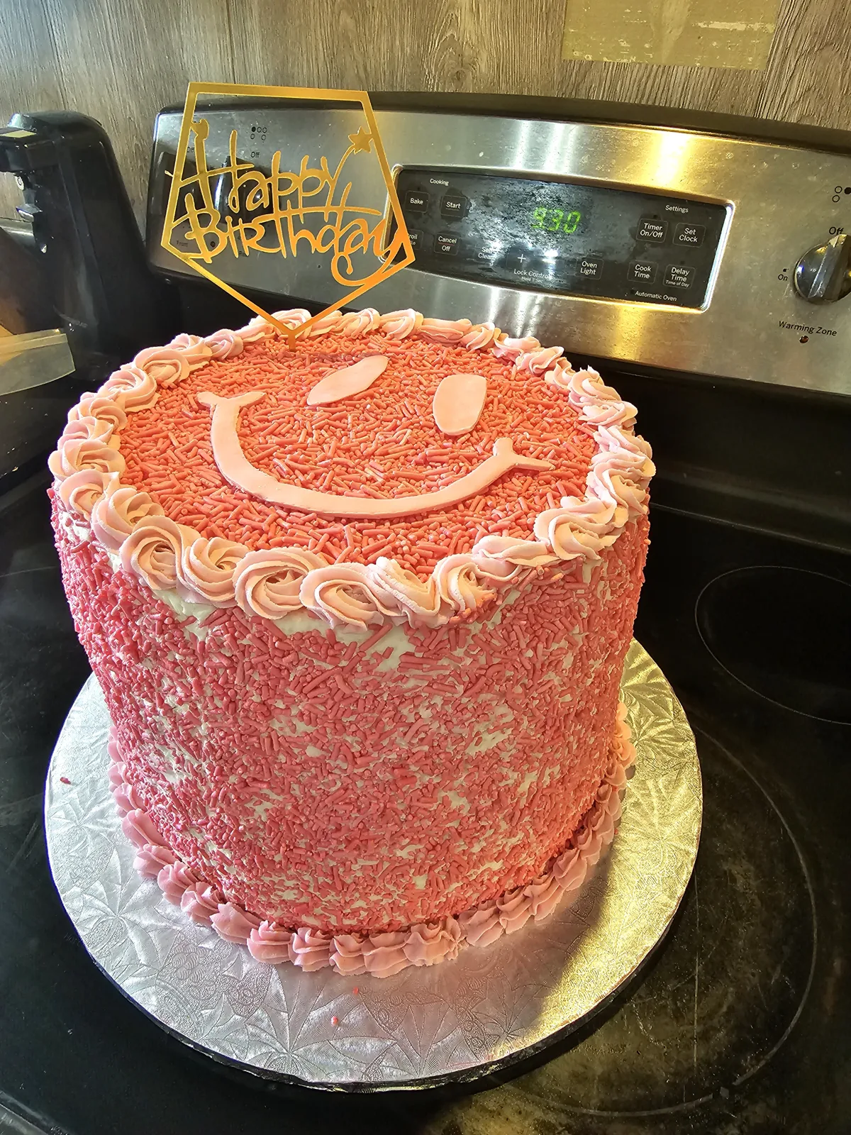 Round birthday cake covered in pink sprinkles with white frosting rosettes and a smiley face design, topped with a gold Happy Birthday topper.
