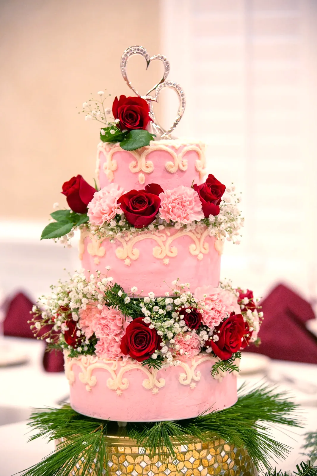 Three tier pink wedding cake featuring white scrollwork piping, layered red roses pink carnations and baby's breath, topped with a silver double heart ornament.