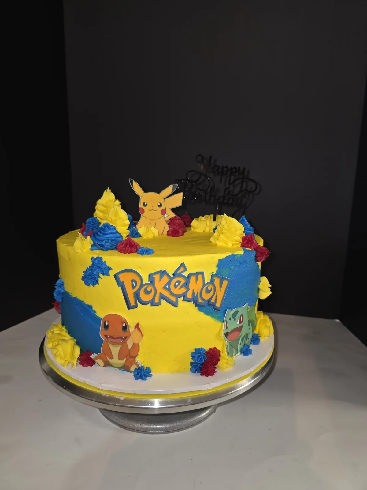 Round yellow and blue frosted birthday cake decorated with Pikachu, Charmander, and Bulbasaur character cutouts plus the Pokemon logo.