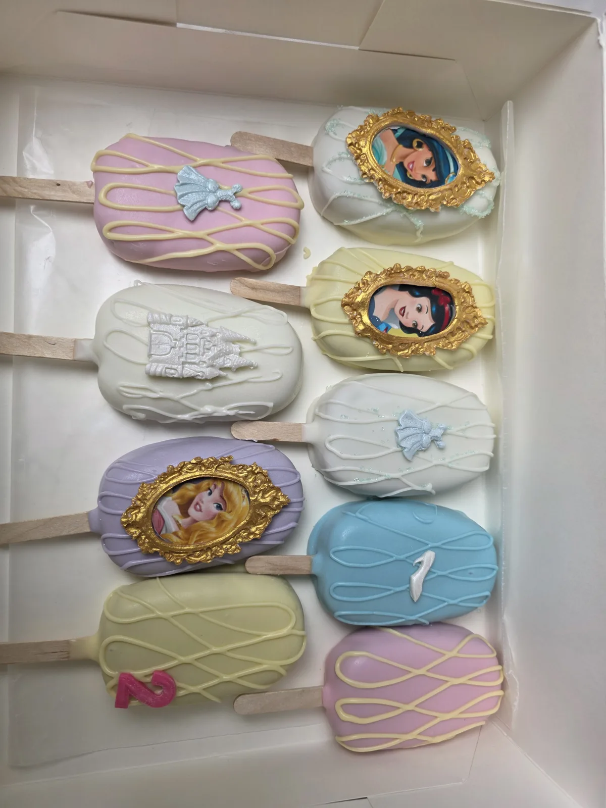 Assorted pastel cakesicles in pink, white, purple, yellow, and blue with white drizzle, featuring princess portraits in gold frames, silver castles, bows, and numbers.