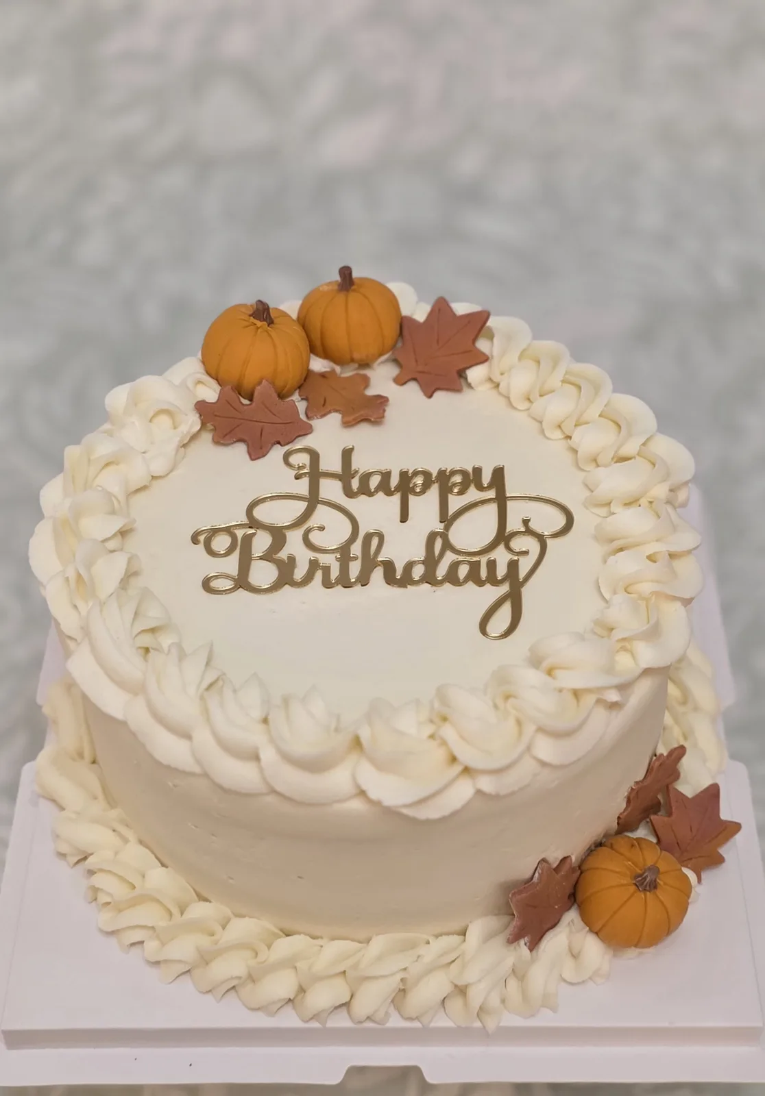 Round cream frosted birthday cake with piped borders, gold Happy Birthday topper, small orange pumpkins and autumn colored fondant leaves.