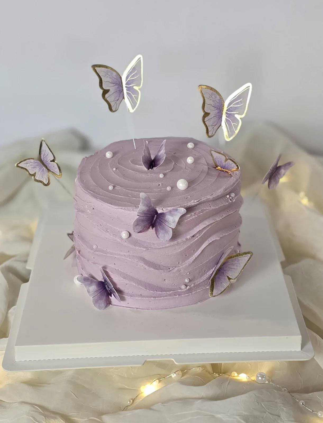 Single tier round lavender buttercream cake decorated with white pearls and purple butterfly accents featuring gold edged toppers.