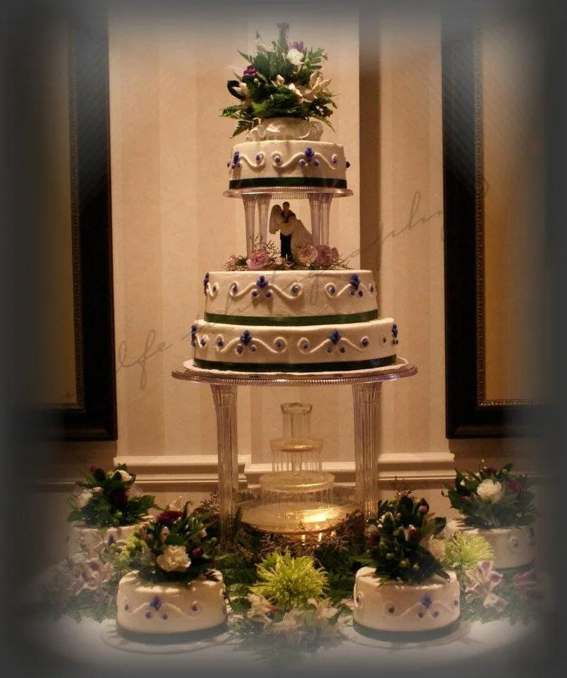 Tall white tiered cake with purple piping, green ribbon trim, floral topper, and matching small cakes surrounding an illuminated fountain base.