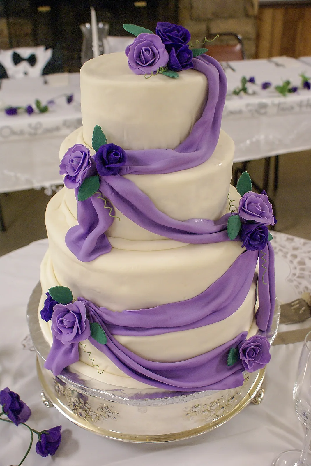 Three tiered white cake draped in cascading purple fondant swags adorned with purple sugar roses, green leaves, and delicate tendrils on a silver stand.
