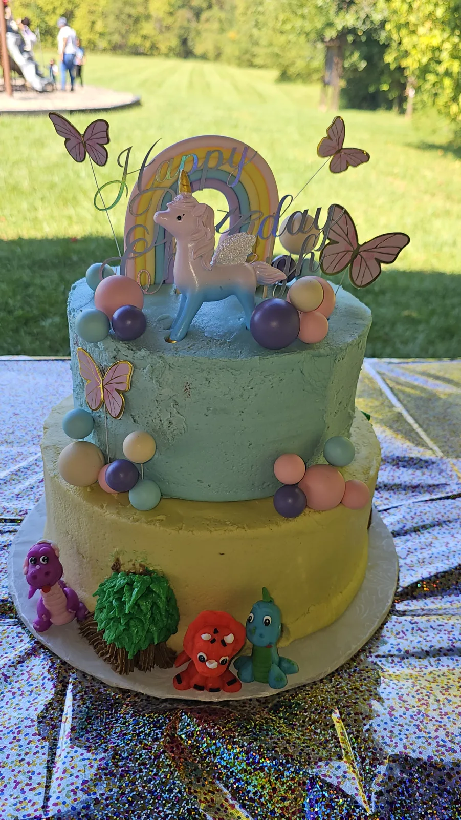 Two tier birthday cake with textured teal and yellow frosting, topped with a pastel rainbow, unicorn figurine, butterfly cutouts, and colorful fondant spheres, with small dinosaur figures around the base.
