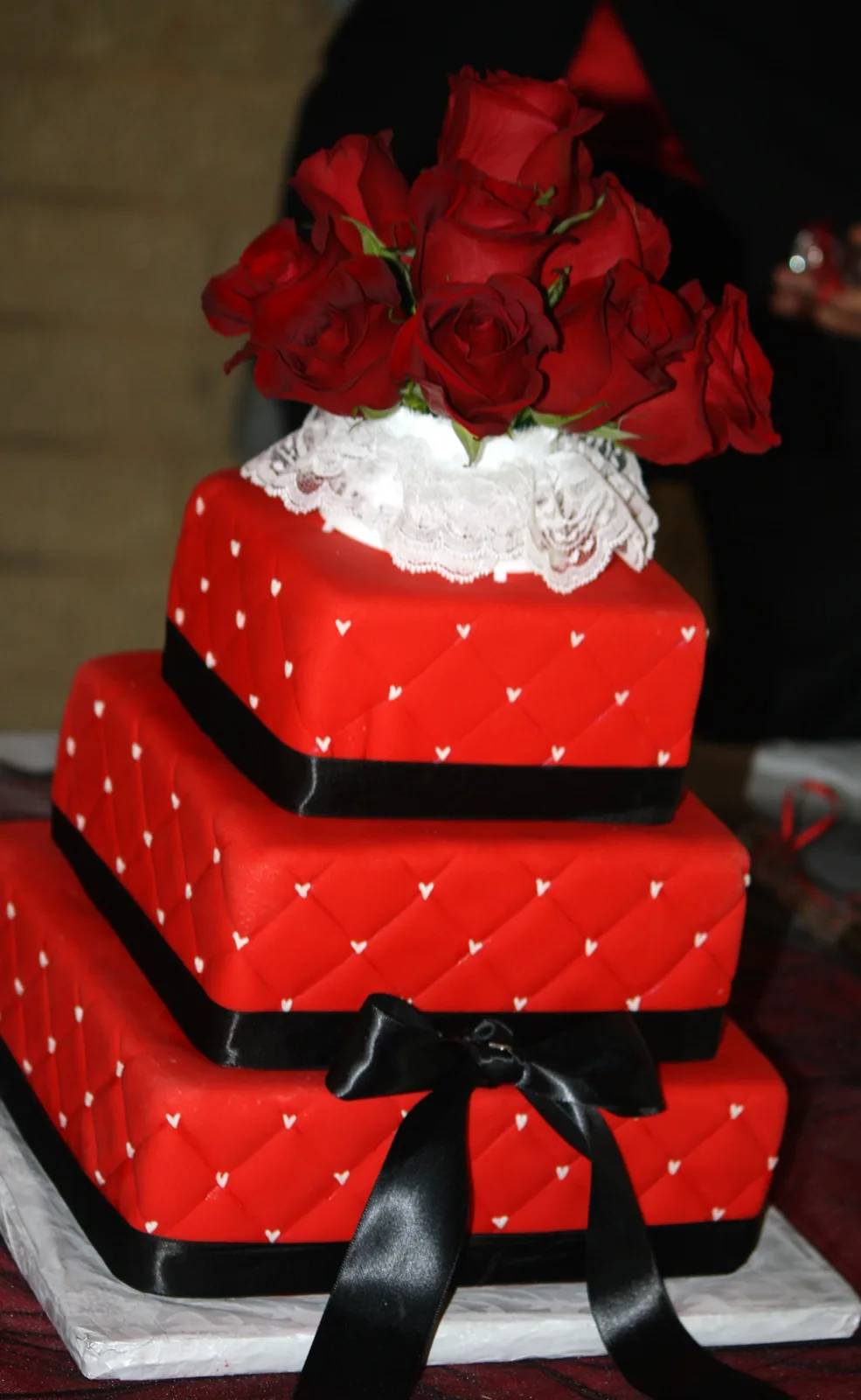 Three square red tiers decorated with white hearts and quilted patterns, accented with black satin ribbons and a large bow, topped with red roses on white lace.