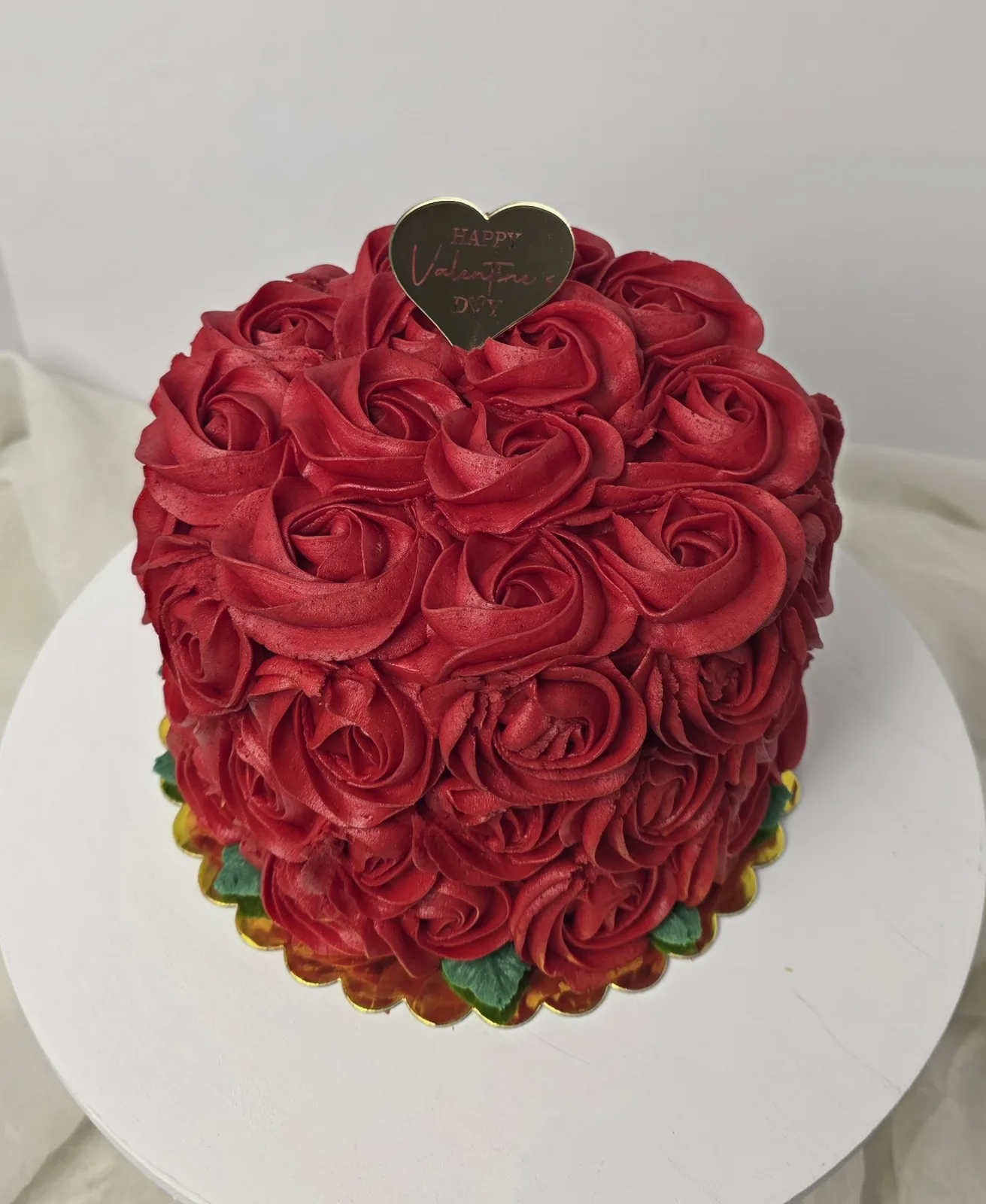 Round cake covered entirely in deep red frosting piped into detailed rose blooms with green leaves at the base, topped with a gold heart topper reading Happy Valentine's Day.