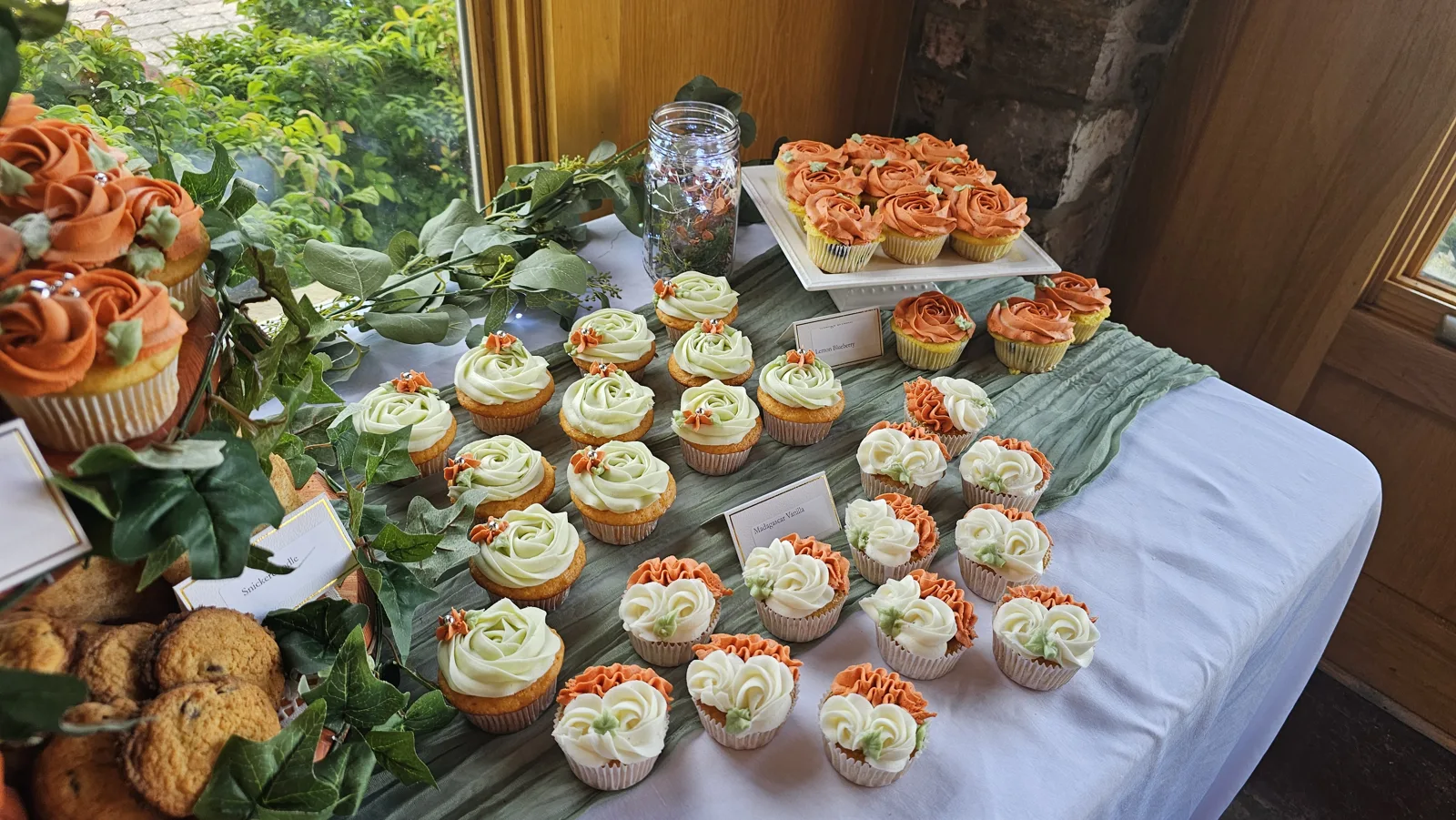 Cupcakes decorated with orange, sage green, and white buttercream roses arranged on a dessert table accented with greenery, cookies, and a tiered rose cake.