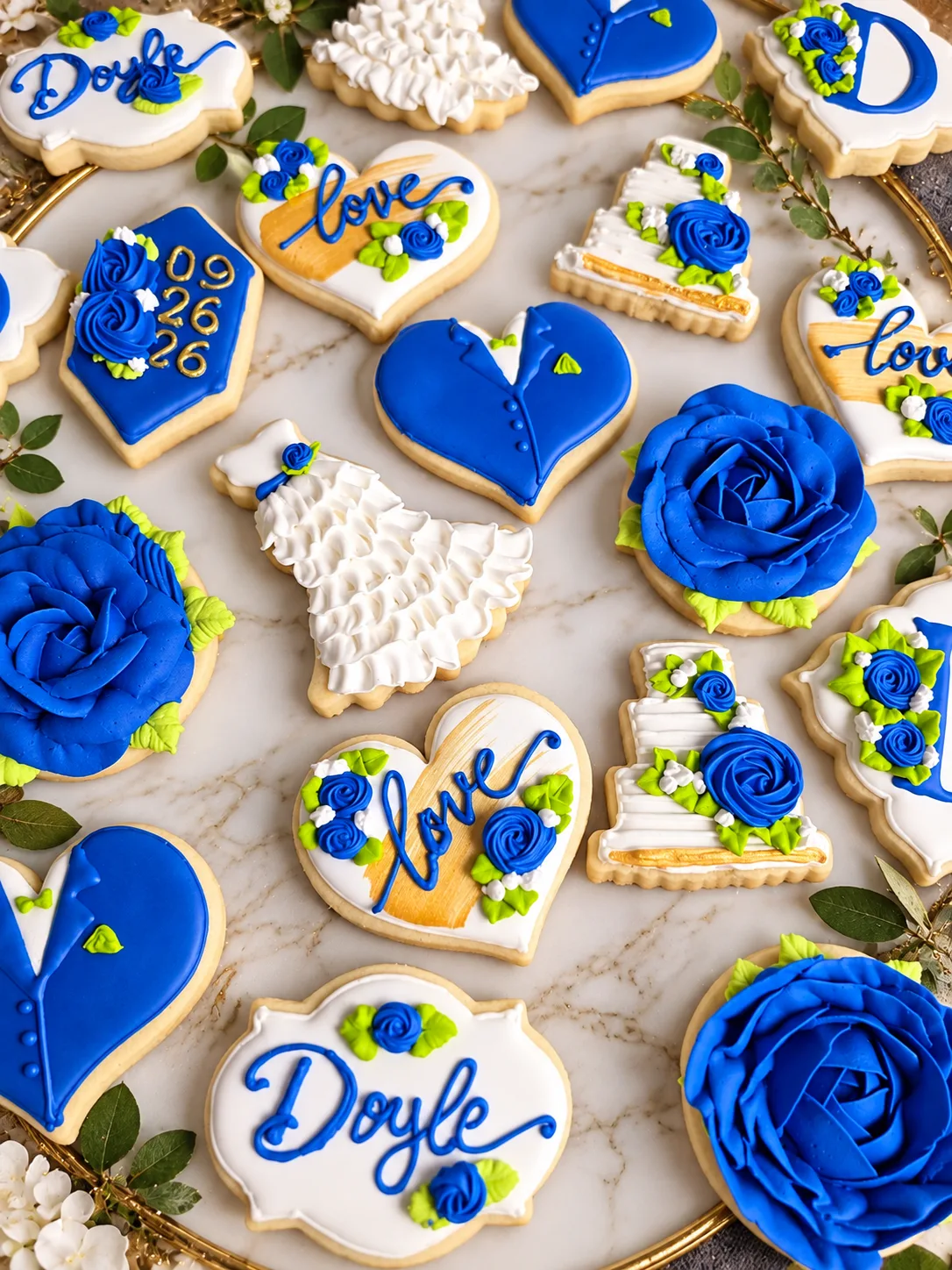 Decorated wedding sugar cookies in royal blue and white, including rose cookies, a ruffled wedding dress, tuxedo hearts, tiered cake cookies, love hearts and name plaques.