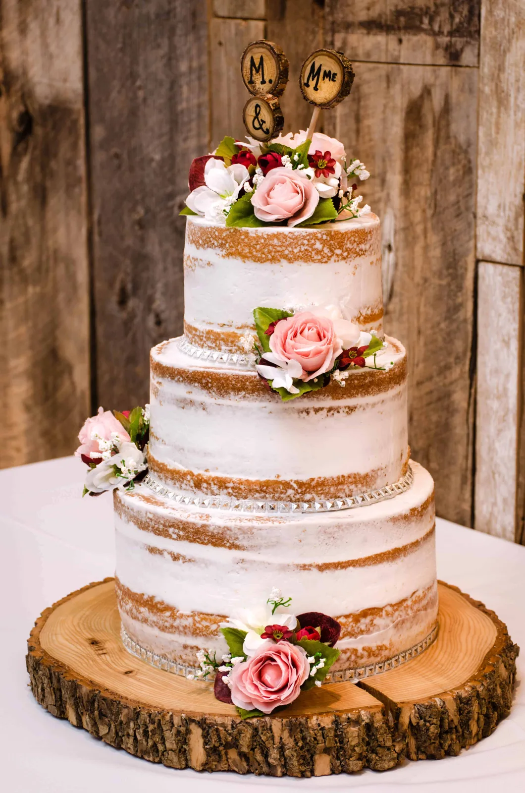 Three tier semi naked wedding cake with white frosting, pink roses, burgundy blooms, and silver ribbon trim, displayed on a wooden log slice base.
