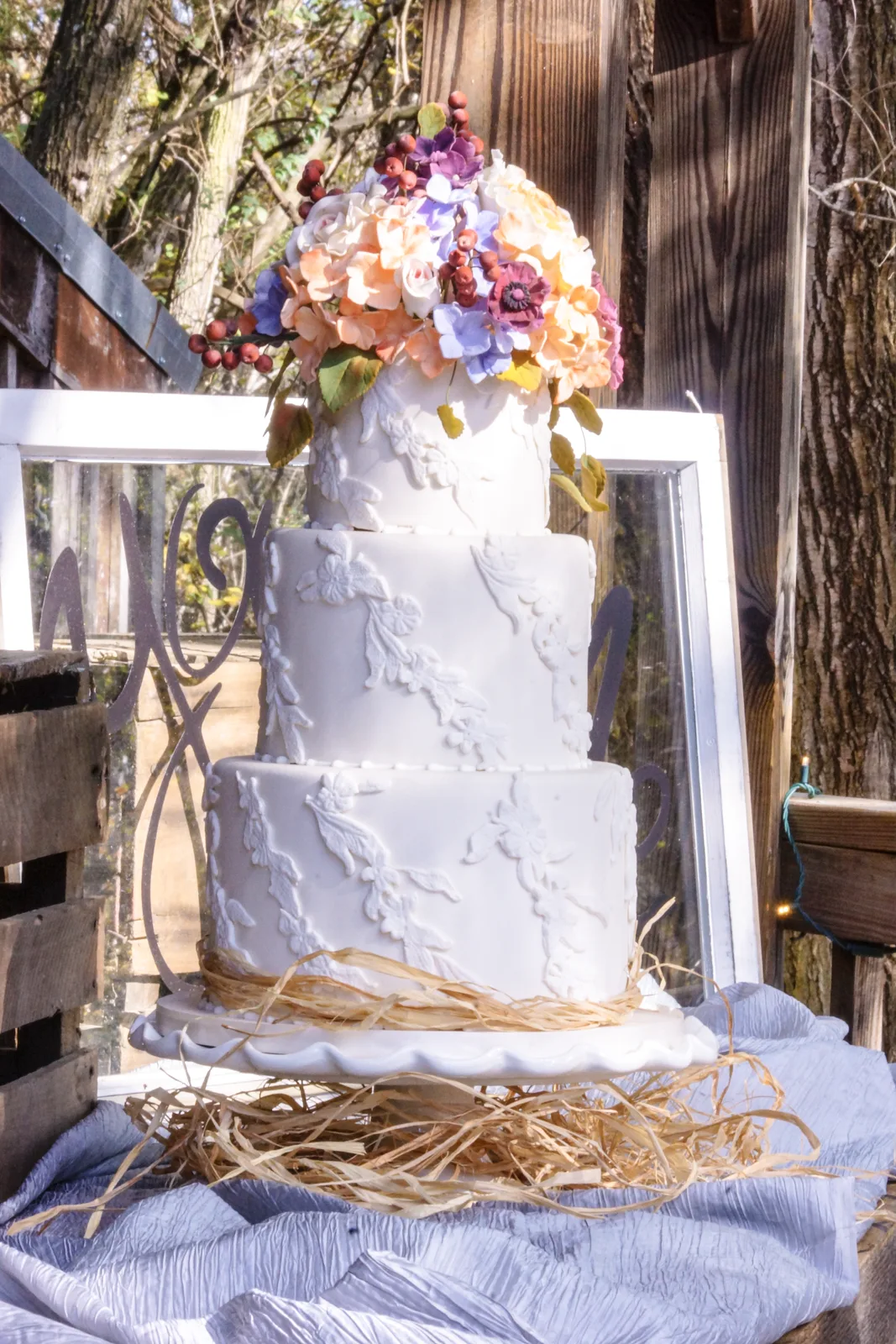 Three tiered white wedding cake with delicate piped floral and leaf motifs, topped with a vibrant sugar flower bouquet of peach roses, purple blooms, and red berries.