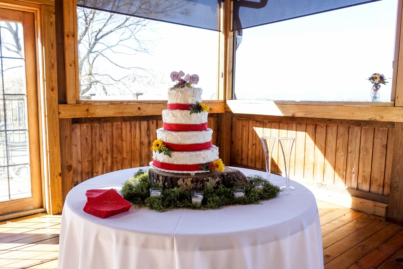 Four tier white buttercream wedding cake with red ribbon trim, sunflower accents, and a Mr and Mrs topper displayed on a tree stump base.