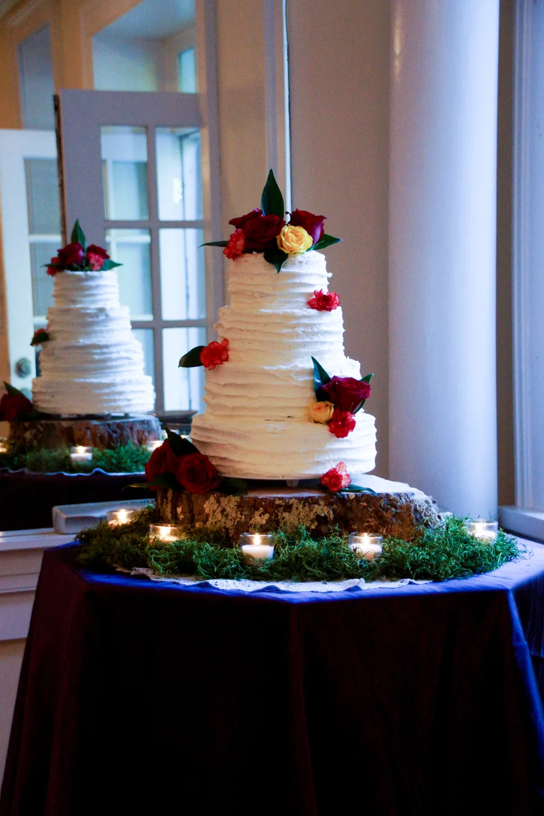 Two tiered white textured buttercream wedding cakes adorned with red and yellow roses, displayed on wood slices with moss and candles.