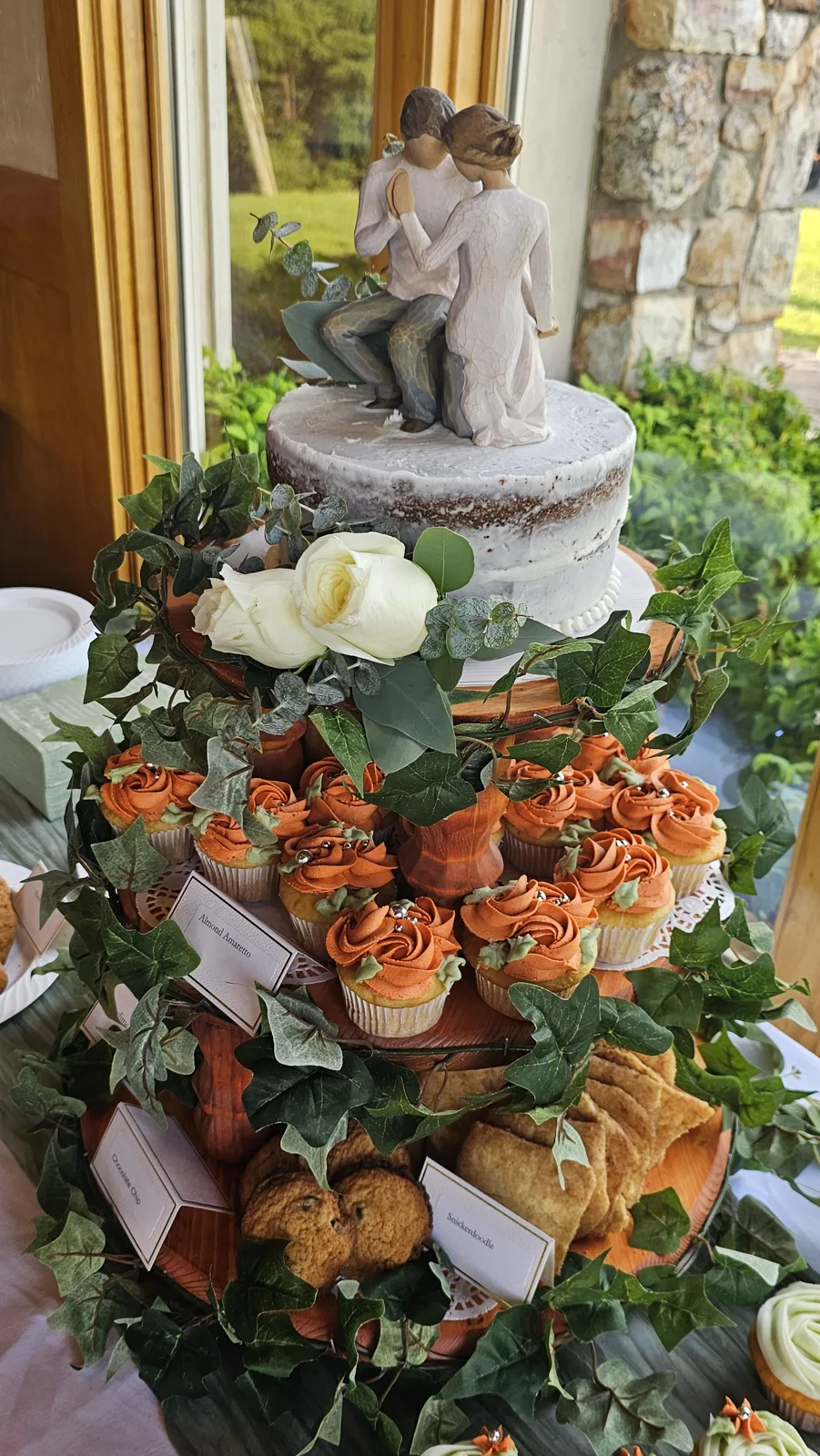 Tiered wooden cake stand with a naked frosted cake topped by a bride and groom figurine, surrounded by ivy garlands, orange frosted cupcakes, and assorted cookies with flavor labels.