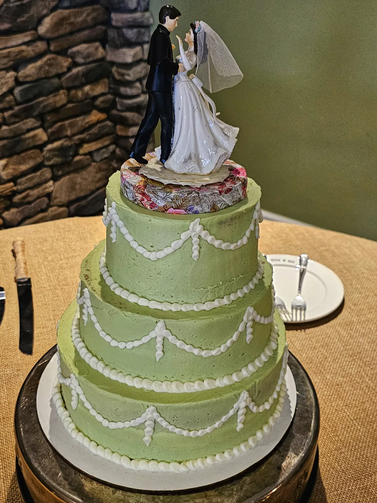 Three tiered sage green wedding cake with white piped garland swags, topped with a bride and groom figurine on a floral patterned base.