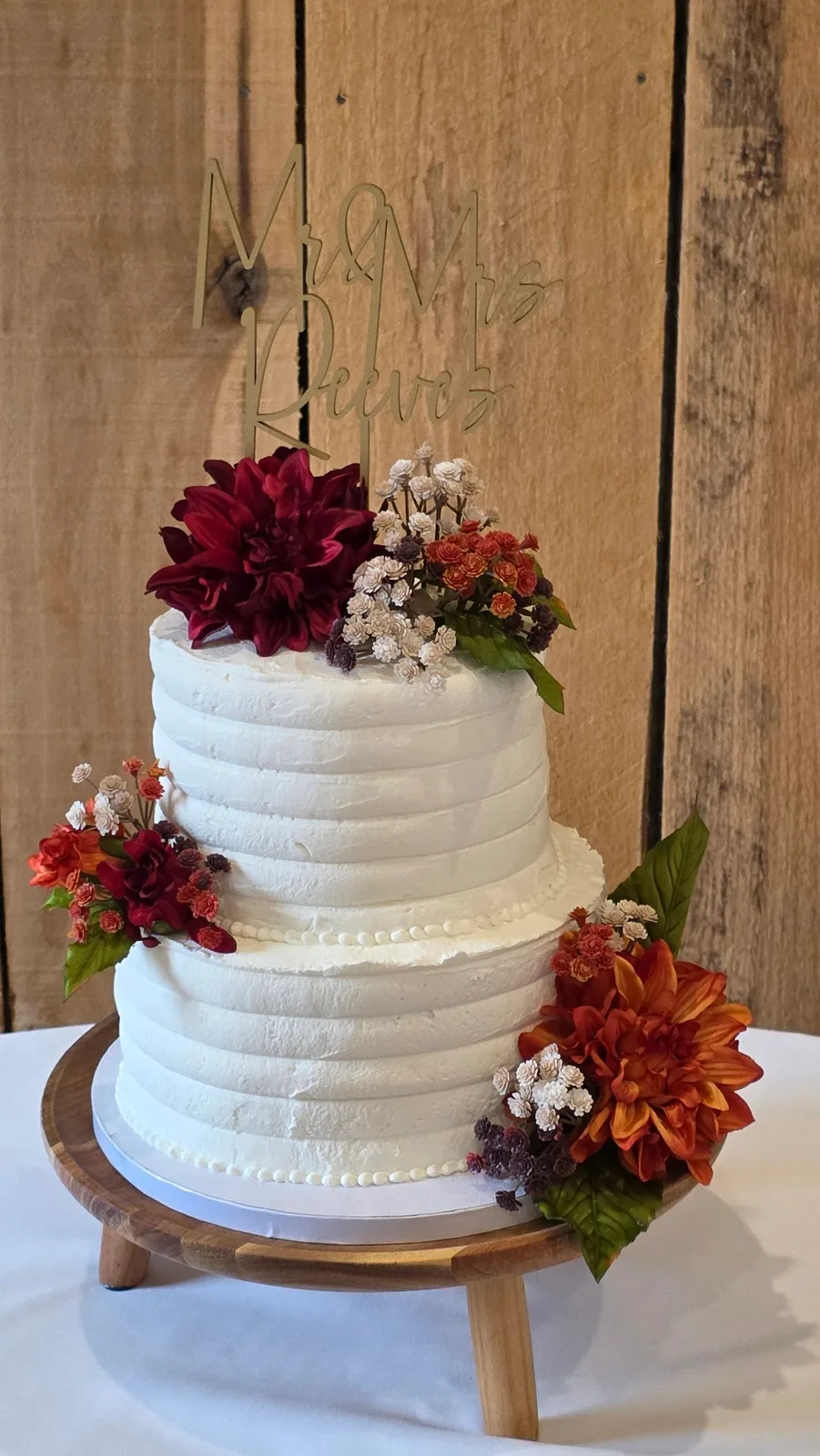 Two-tier white textured wedding cake with gold Mr & Mrs Reeves topper, burgundy and rust dahlias, on a wooden stand.