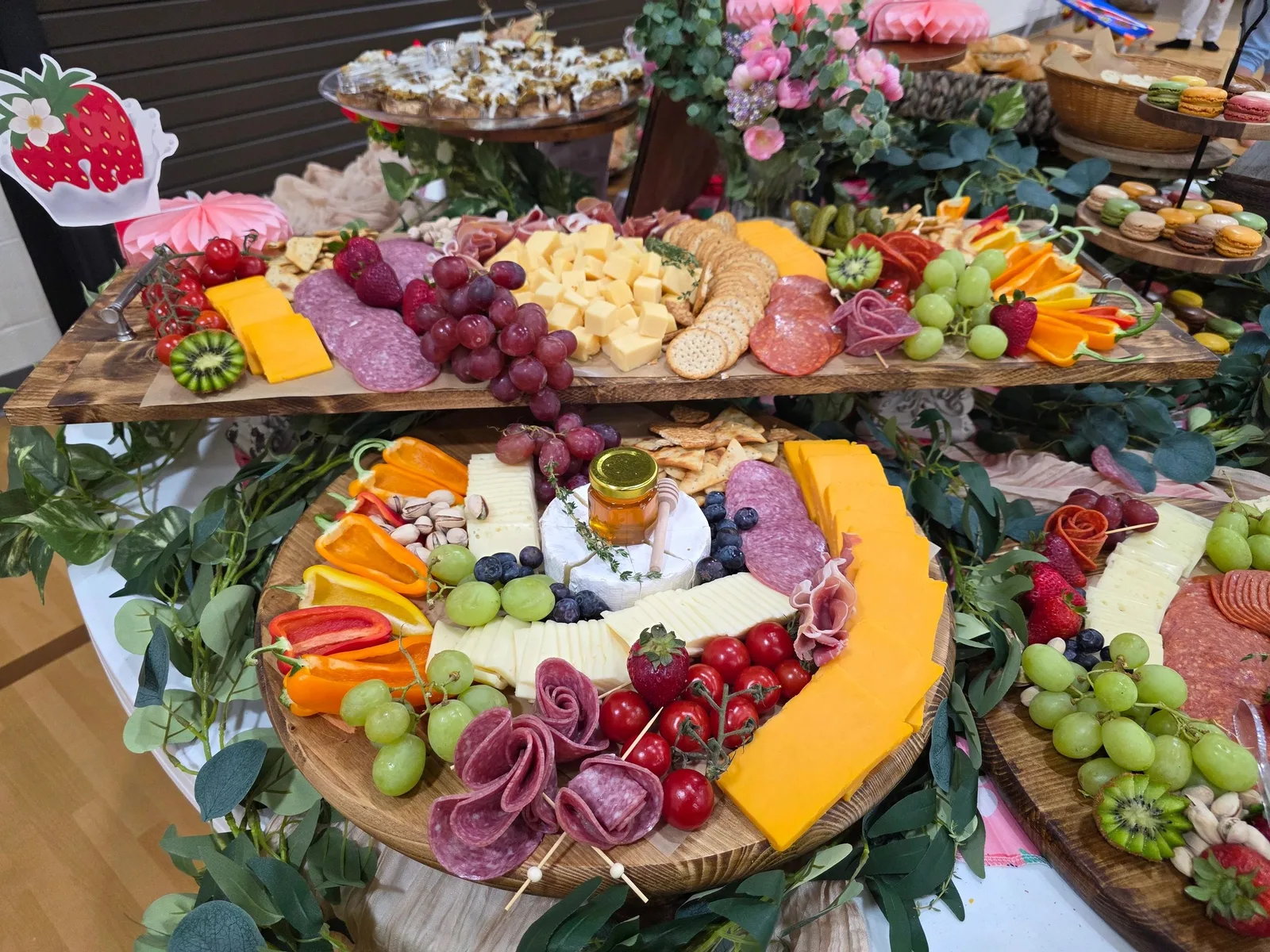 Lavish charcuterie spread with cheeses, salami, grapes, strawberries, peppers, and honey, surrounded by greenery and macarons.