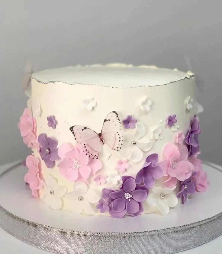 A smooth white cake sits on a silver-edged board, its base clustered with pink, lavender, and white flowers featuring silver centers.