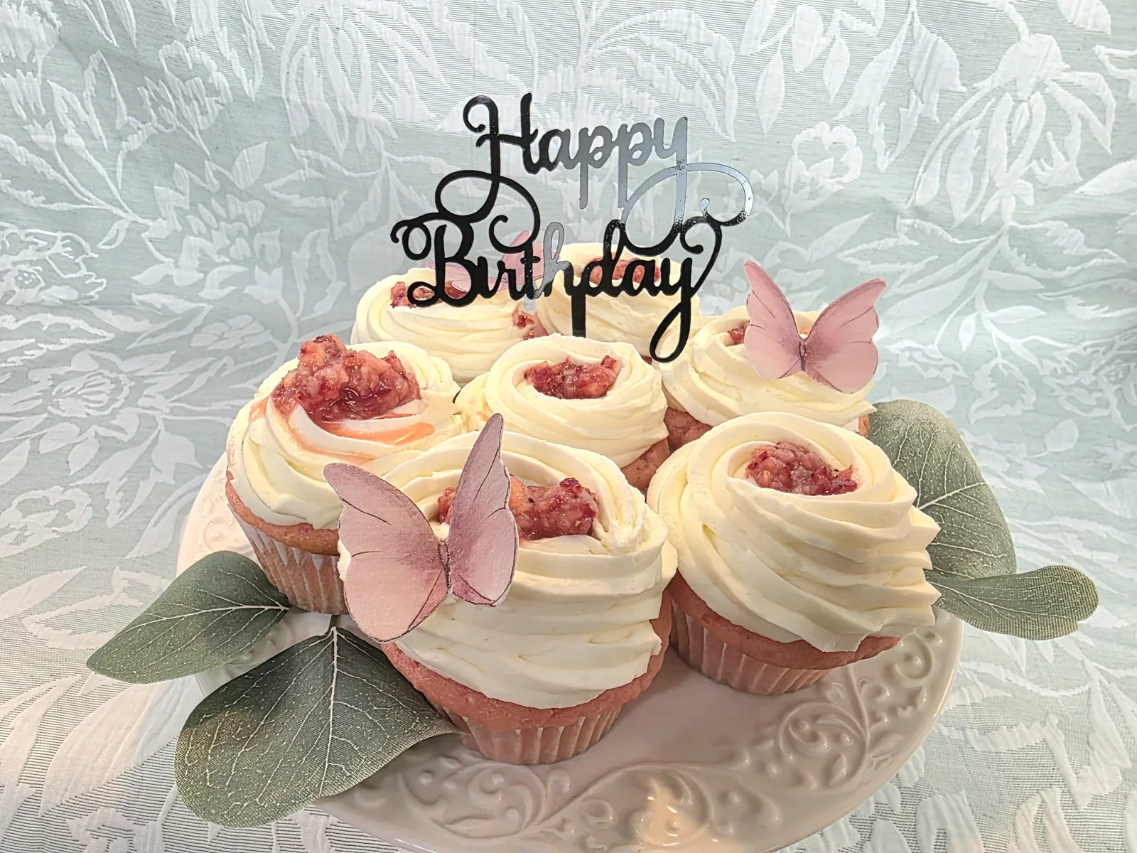 Pink and white swirled cupcakes topped with strawberry filling, pink butterfly decorations, and a Happy Birthday topper on a white pedestal stand with green leaves.