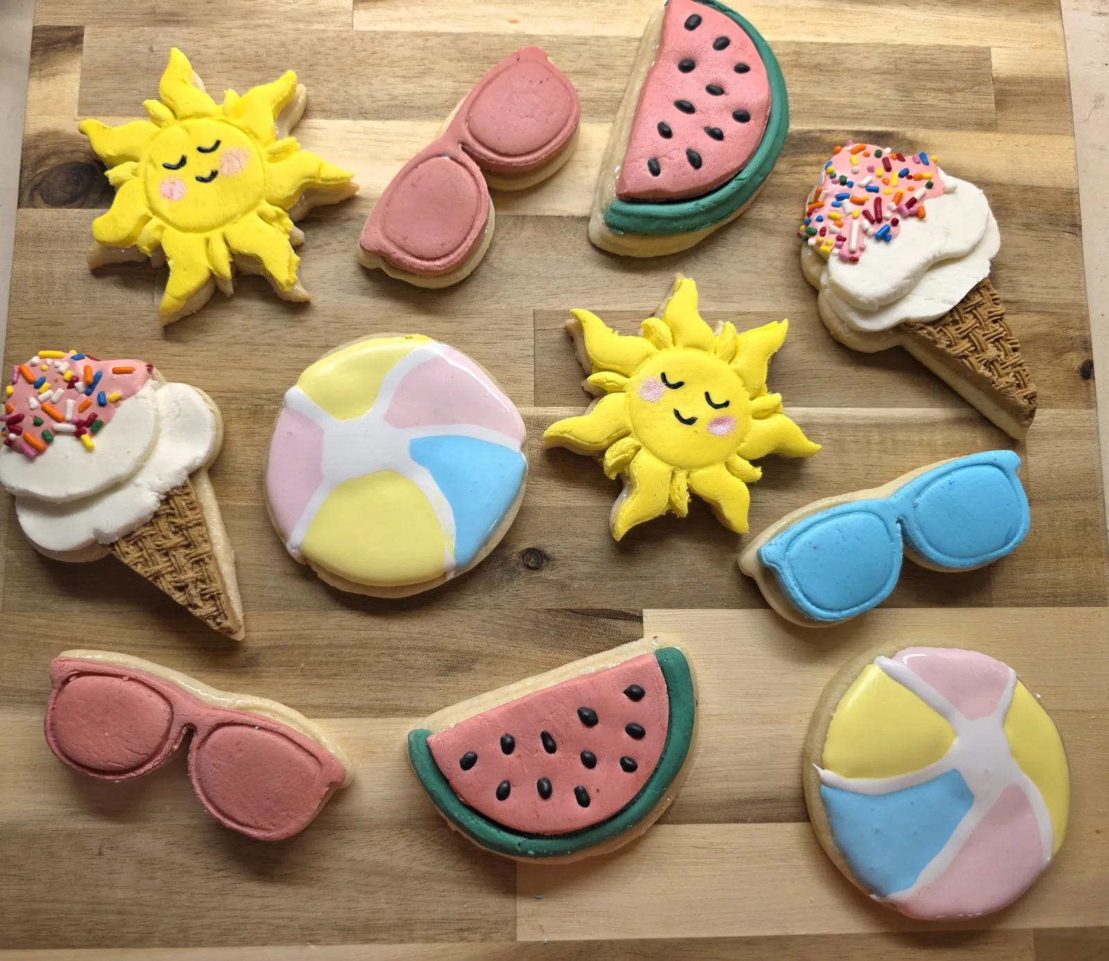 Assortment of decorated sugar cookies featuring yellow suns with closed eyes, pink and blue sunglasses, watermelon slices, pastel beach balls, and ice cream cones with rainbow sprinkles.