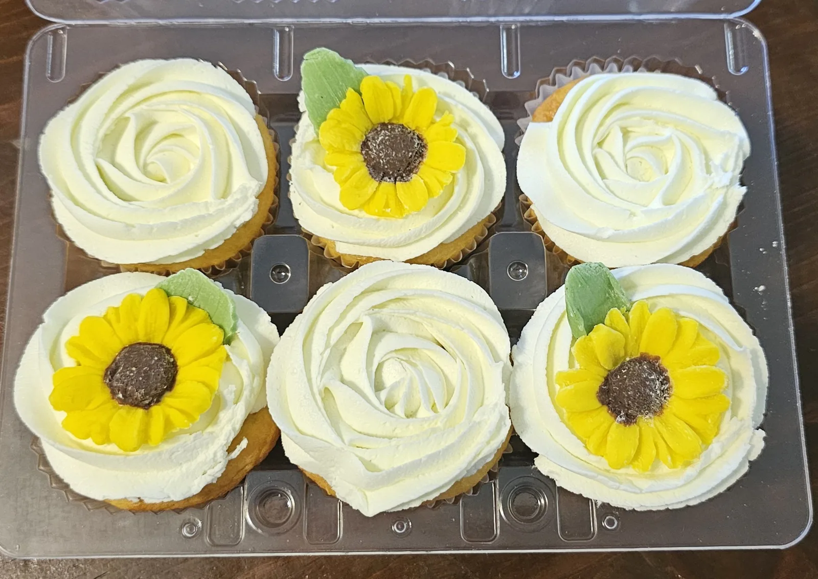 Six cupcakes in a clear container topped with white frosting swirls, three of them decorated with yellow sunflower toppers featuring brown centers and green leaves.