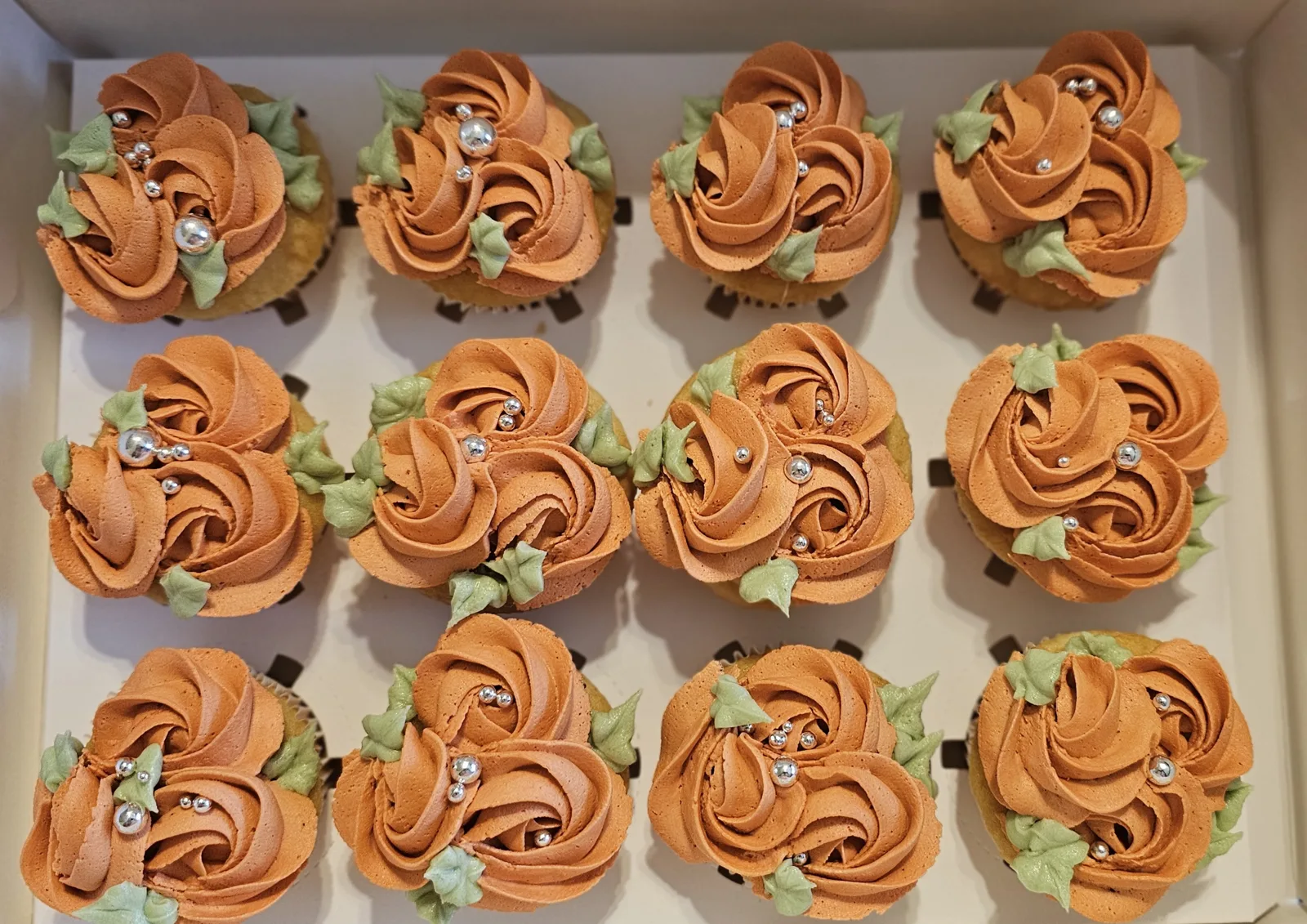 Twelve cupcakes with terracotta orange rose piped frosting, sage green leaves, and silver sugar pearls arranged in a white box.