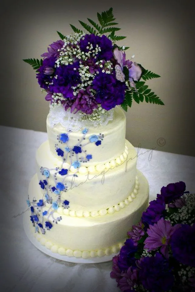 Three tier smooth white wedding cake decorated with blue and purple piped sugar flowers cascading down the side and topped with purple chrysanthemums, baby's breath, and green ferns.