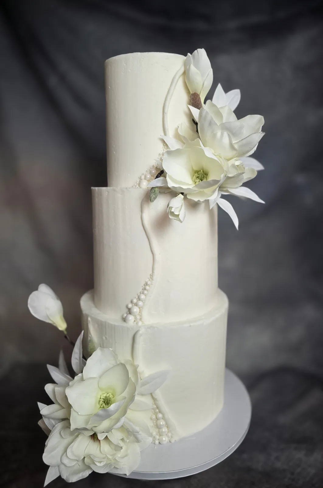 Tall three tiered white wedding cake decorated with large sugar magnolia blossoms, white peonies, and cascading strands of sugar pearls against a dark background.