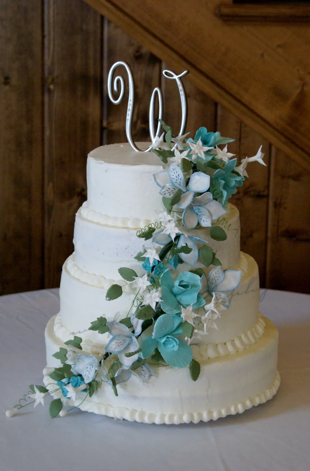 Four tier white wedding cake decorated with cascading turquoise and white sugar flowers, green leaves, and pearl borders beneath a silver wire monogram topper.