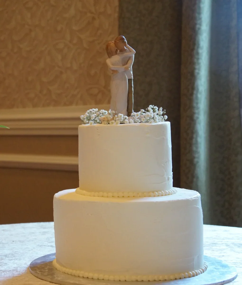 Two-tier white buttercream wedding cake with a bride and groom topper and baby's breath, cropped close so the cake is the focus.
