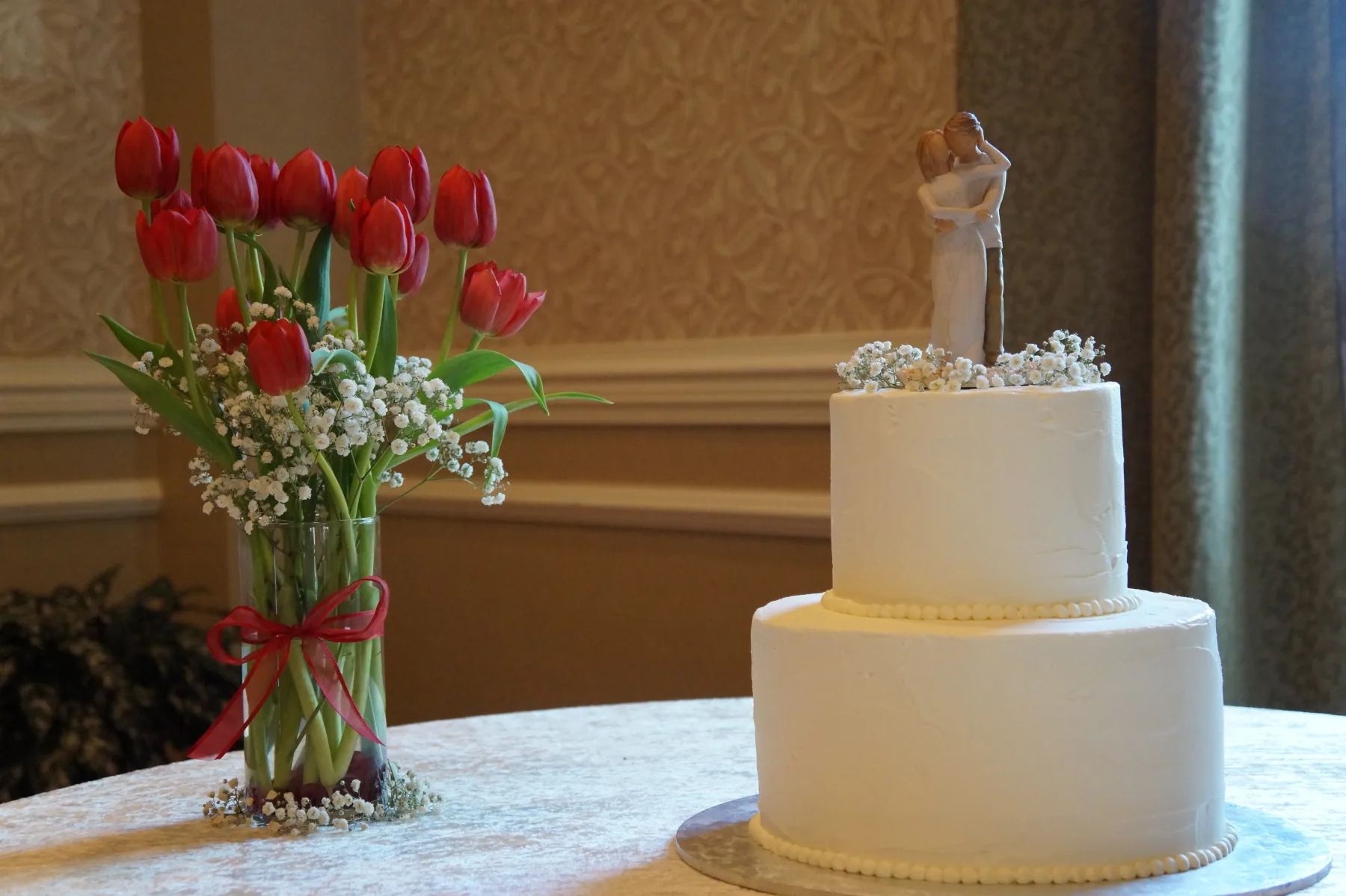 Two-tier white buttercream wedding cake with a bride and groom topper and baby’s breath, beside a glass vase of red tulips.