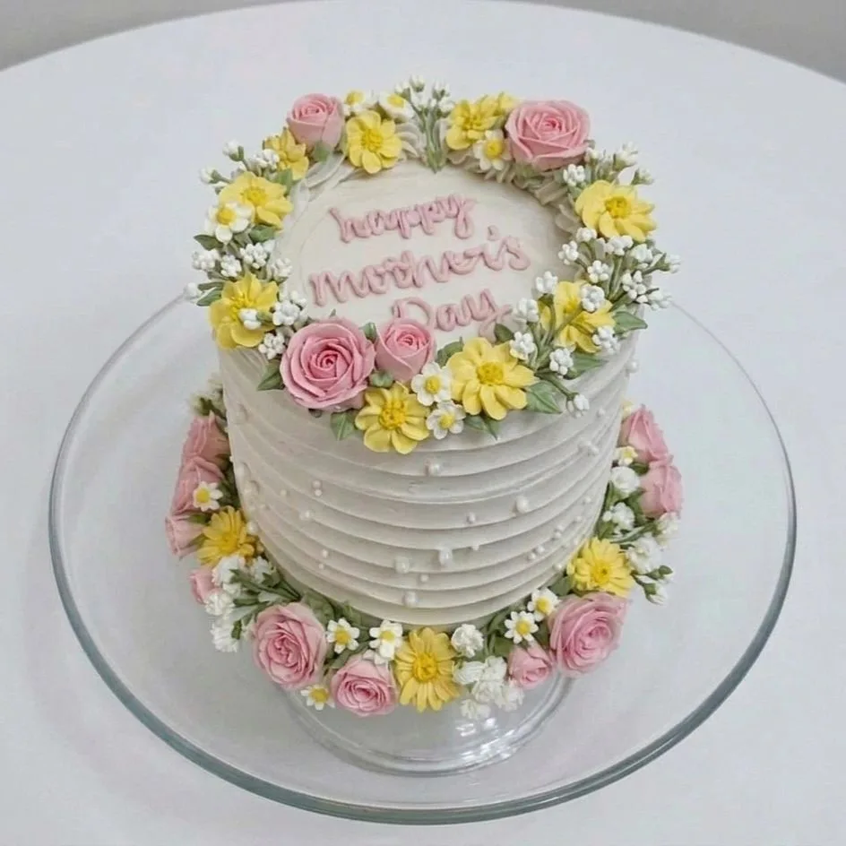 White frosted round cake on a glass stand, decorated with pink and yellow buttercream flowers and "Happy Mother's Day" written in pink script on top.